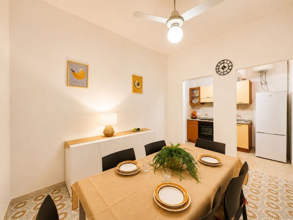 Uma sala de jantar com uma mesa posta para quatro pessoas, um ventilador de teto e um relógio na parede.