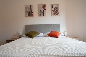 Uma cama com cobertura branca texturizada, duas almofadas, cabeceira cinza e três obras de arte emolduradas na parede.