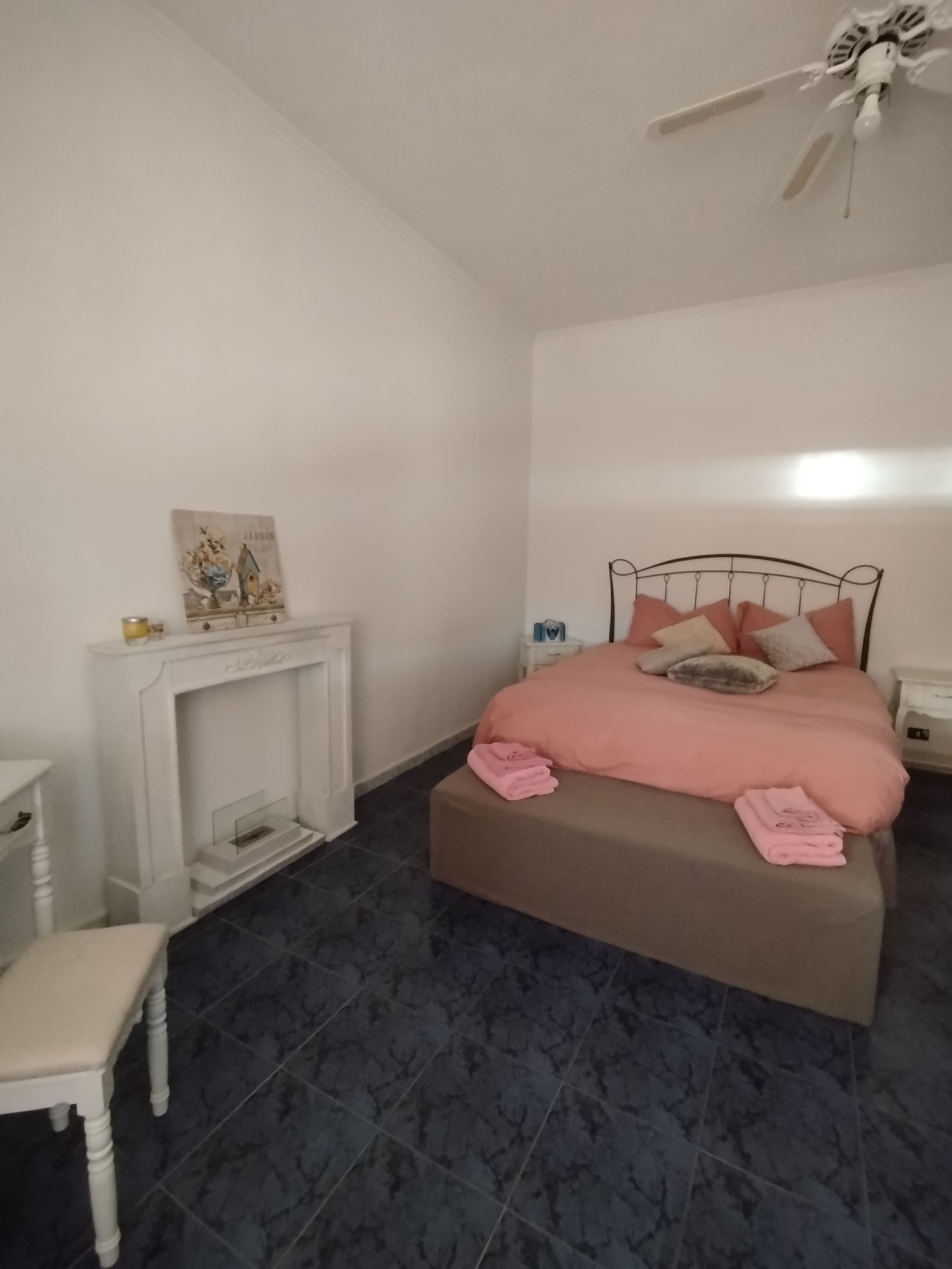 Une chambre à coucher avec un lit, une table de chevet, une chaise blanche, une cheminée et un ventilateur de plafond.