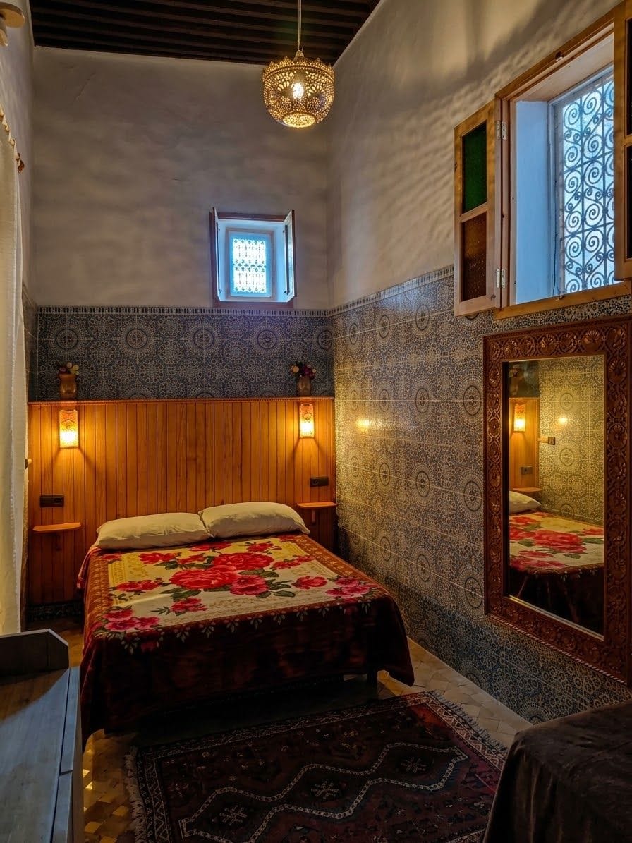 Une chambre à coucher avec un lit, une fenêtre ornée de fer forgé, un luminaire fixé au mur et un tapis à motifs.