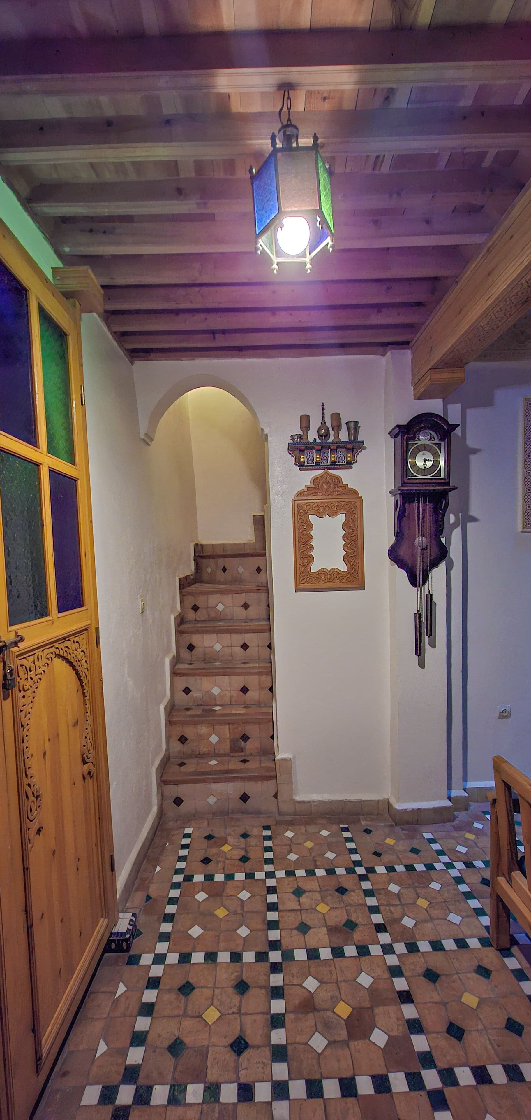 Un escalier menant à un palier doté d’un sol à motifs, d’une porte en bois, d’une horloge murale et d’un luminaire suspendu.