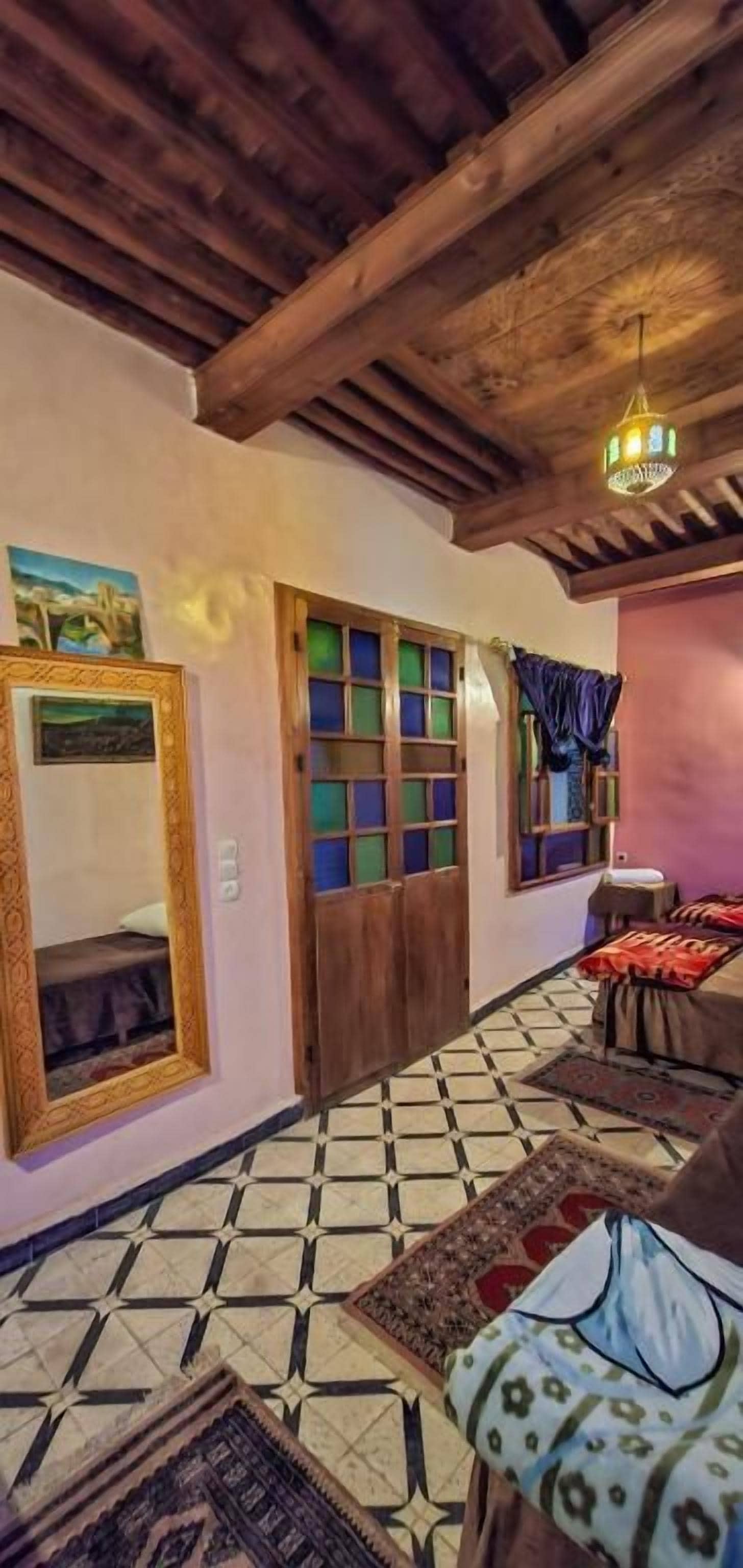 Una habitación con techo de madera, suelo de baldosas, cama con cubrecama estampado, puerta de madera con ventanal y lámpara colgante.