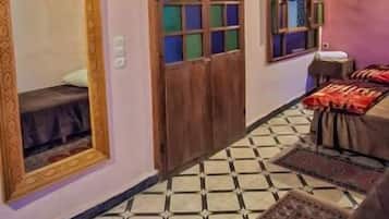 Una habitación con techo de madera, suelo de baldosas, cama con cubrecama estampado, puerta de madera con ventanal y lámpara colgante.