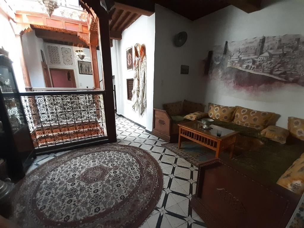 Un salon avec un tapis à motifs, un canapé agrémenté de coussins à motifs, une table basse et un mur orné d’un tableau représentant un paysage urbain.