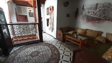 Een woonkamer met een kamerkleed met patroon, een bank met gekleurde kussens, een bijzettafeltje en een wand met een stadsgezicht schilderij.