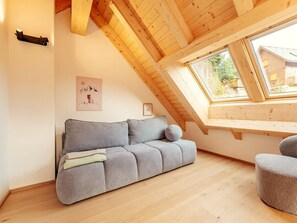 A loft-style room with a wooden ceiling, a grey sofa, a small ottoman, and a window overlooking a residential area. - Chalet # 8 for 2 People With Sauna (Oberwölz)