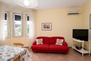 A living room with a red sofa, a television on a stand, a table with a patterned tablecloth, and an air conditioning unit on the wall. - Wonderful  villa for 5 people with A/C, WIFI, TV and patio (Marina Di Modica)