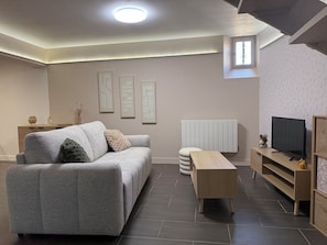 A modern living room with a grey sofa, a wooden coffee table, a TV stand, and a radiator. - L'Oustal Del Nort - Vacation Rental for 6 People Near Millau (Aveyron) (Paulhe)