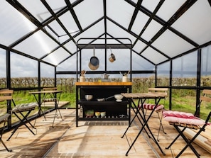 Een kas met een tafel en stoelen, een hangende kruidenrek en uitzicht op het landschap.