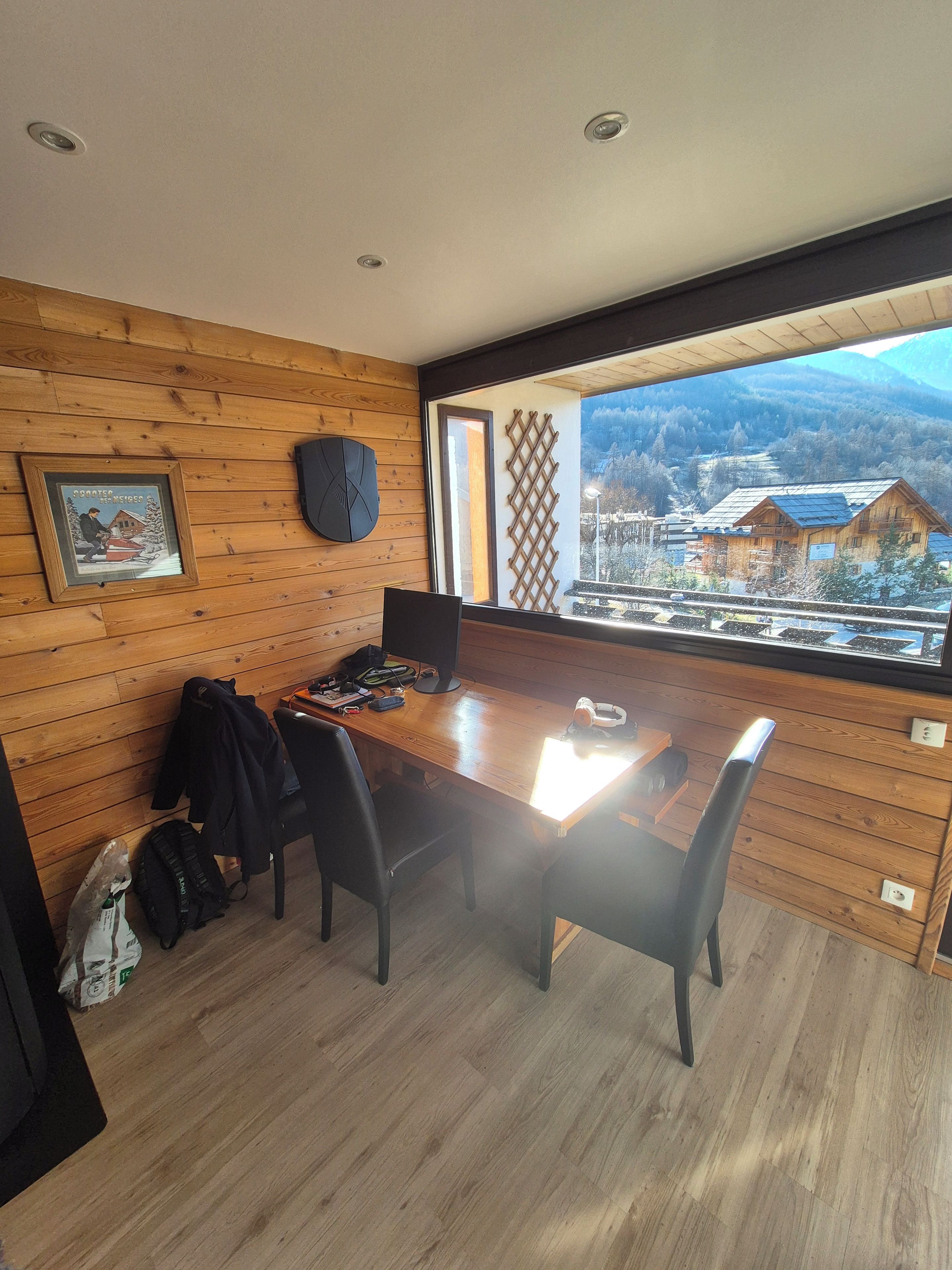 A room with a wooden paneled wall, a dining table with chairs, a framed picture, and a view of a mountainous landscape through a window.
