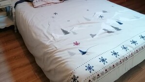 A bedroom with a wooden ceiling, a bed with a white bedspread featuring leaf patterns, two pillows, a nightstand, and a small table. - Vrbo Property (Saint-Chaffrey)