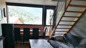 A room with a wooden table, a sofa, a staircase, and a view of a forested area. - Vrbo Property (Saint-Chaffrey)