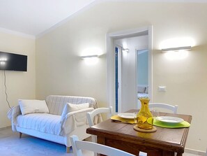 A living room with a sofa, a wooden dining table with chairs, a flat-screen TV mounted on the wall, and a door leading to another room. - Beautiful private villa for 2 people with A/C, WIFI, TV, patio and pets allowed (Montescudaio)