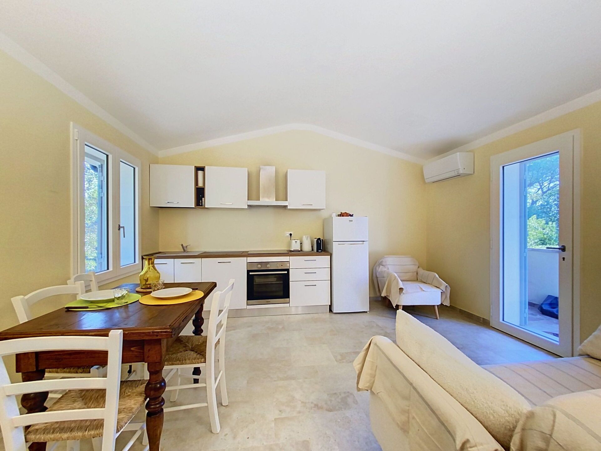 A compact kitchen with white cabinets, a dining table with chairs, a sofa, and a sliding glass door leading to an outdoor area.