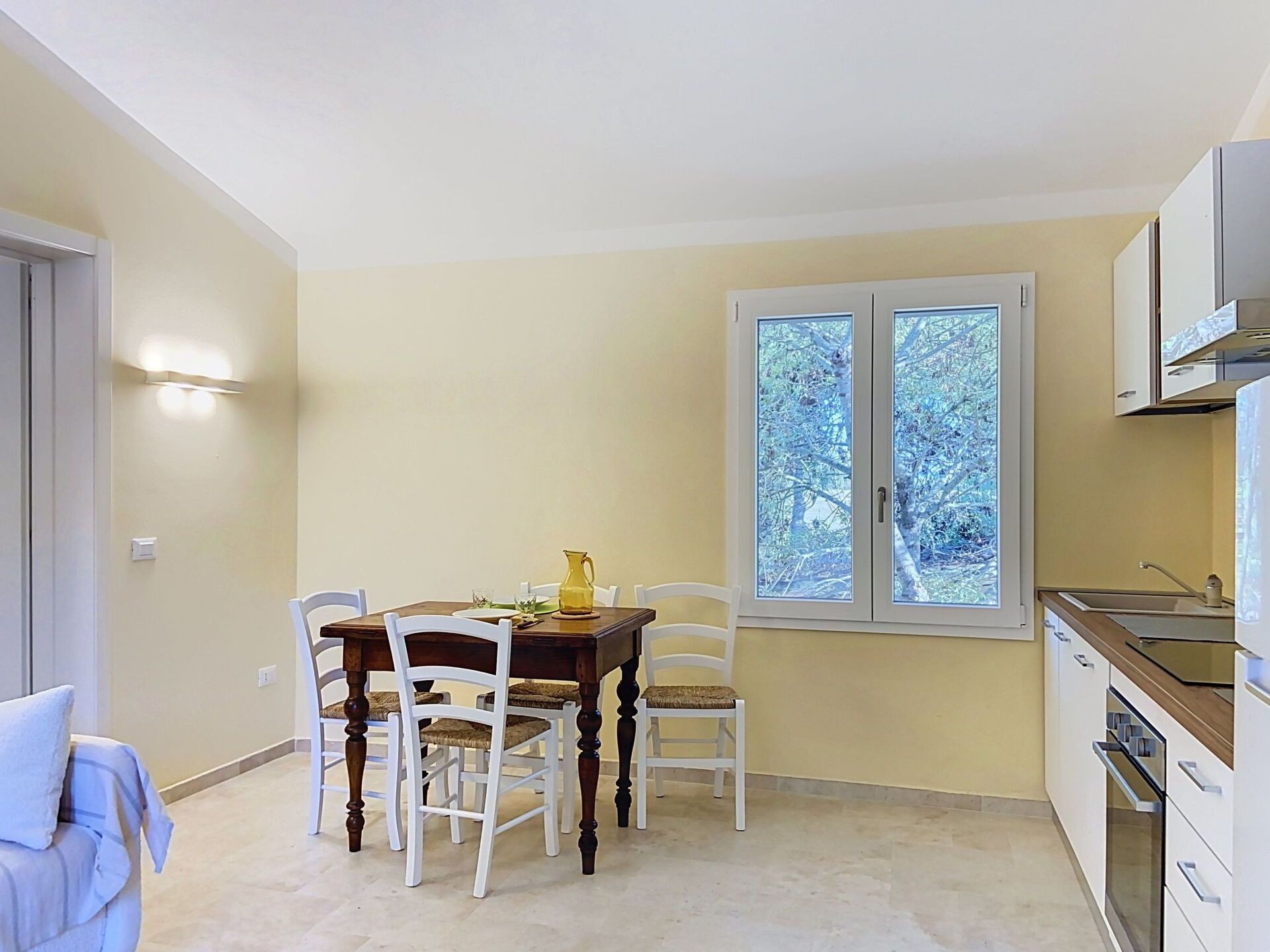 A dining area with a table and chairs, a window with a view of trees, and a kitchen area with cabinets and a sink.