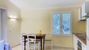 A dining area with a table and chairs, a window with a view of trees, and a kitchen area with cabinets and a sink.