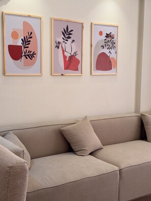 A beige sofa with a cushion, three framed abstract artworks on the wall, and a plain wall. - 3BRs Lightwood Living Aprt (Cairo Governorate)