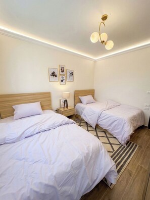 A hotel room with two beds, a bedside table with a lamp, and wall-mounted artwork. - 3BRs Lightwood Living Aprt (Cairo Governorate)