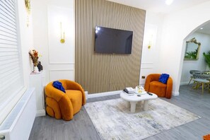 A modern living room with two orange armchairs, a white coffee table, and a flat-screen TV mounted on a wooden panel wall. - Cosy 3 Bedroom House In Manchester Sleeps 7 (Salford)