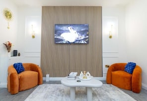 A living room with a television mounted on a wall, two orange armchairs with blue cushions, a white coffee table, and a decorative wall panel. - Cosy 3 Bedroom House In Manchester Sleeps 7 (Salford)