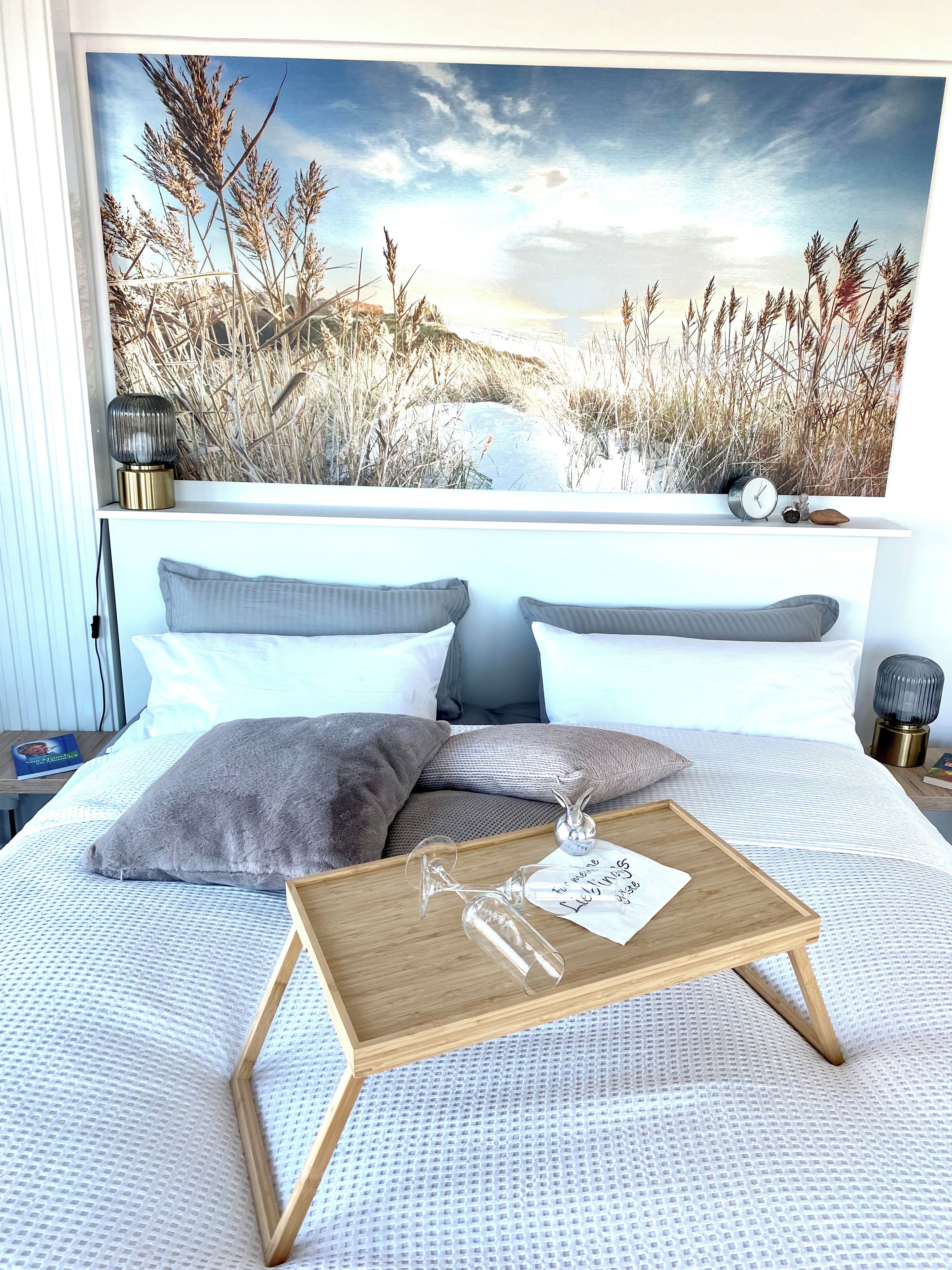 A bedroom with a large window, a bed with white bedding, a bedside table with a lamp, and a wall-mounted clock.