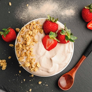 Eine Schüssel Joghurt mit Granola und Erdbeeren auf einer dunklen Unterlage.