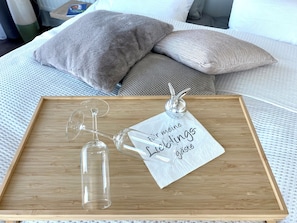 Ein Holztablett mit einer klaren Glasflasche und einem Weinglas auf einem Bett mit Kissen.