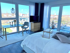 Ein Hotelzimmer mit Bett, Schreibtisch, Stuhl, Fernseher und großen Fenstern mit Blick auf die Stadt.
