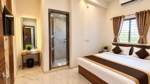 A hotel room with a bed, a mirror, a small table with a plant, a trash bin, and a window with curtains. - Shree Radhe Shyam Palace (Khatoo)