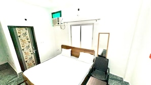 A single-bed room with a wooden headboard, a small table, a chair, a window, and a wall-mounted air conditioning unit. - Namo Krishn Stay Kunj (Mathura)