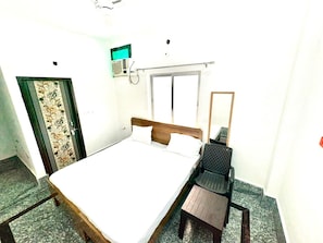 A single-bed room with a wooden headboard, a small table, a chair, a window, and a wall-mounted air conditioning unit. - Namo Krishn Stay Kunj (Mathura)