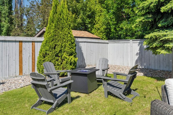 A backyard with a wooden deck, a tall coniferous tree, and outdoor furniture including two Adirondack chairs and a square table.
