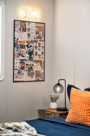 Ein Schlafzimmer mit einem an der Wand befestigten Brett voller Hockey-Erinnerungsstücke, einem Nachttisch mit Lampe und einem Bett mit blauer Decke und orangefarbenem Kissen.
