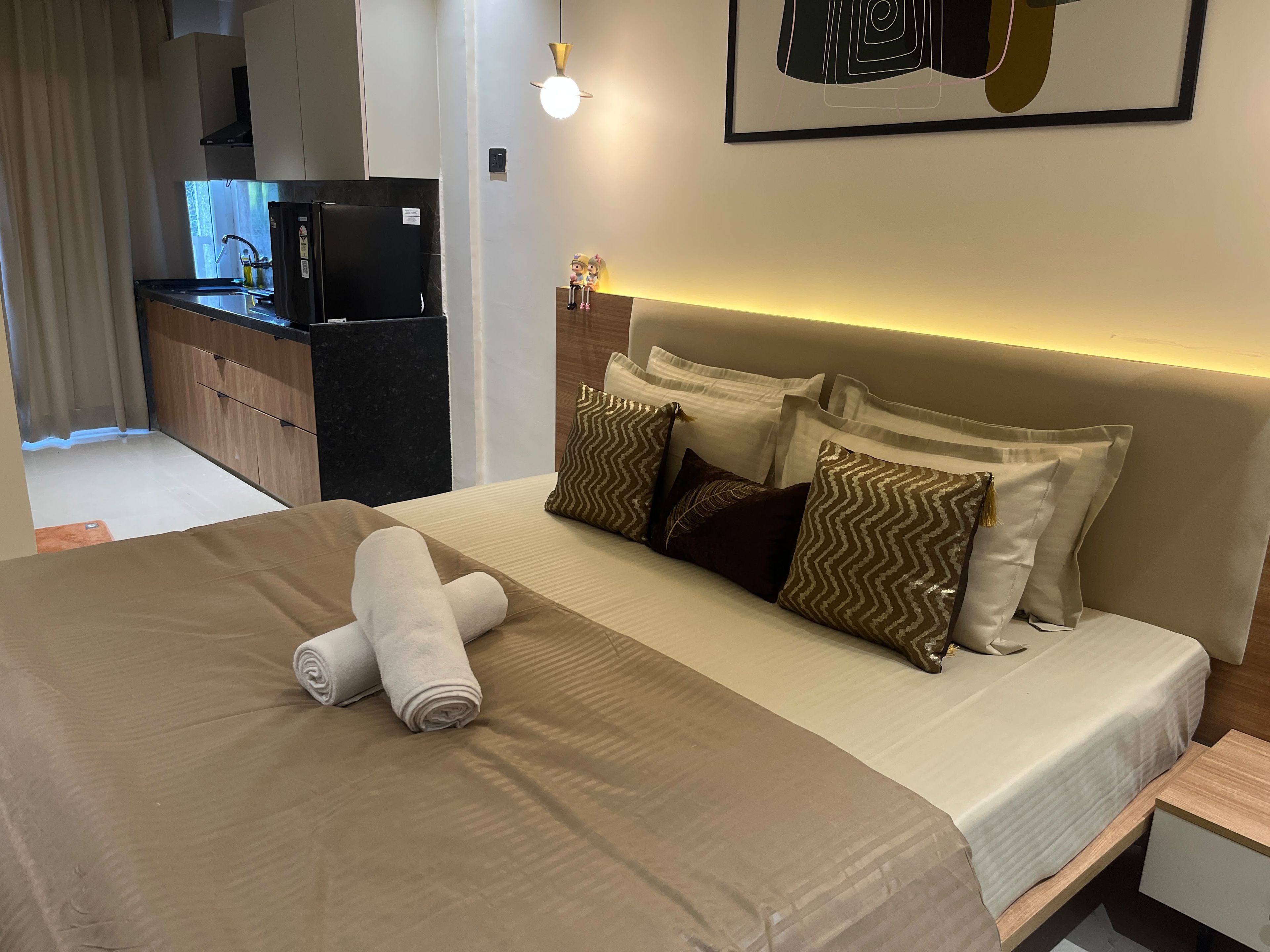 Une chambre d’hôtel avec un lit, des oreillers, une table de chevet, une commode, une œuvre d’art accrochée au mur et, au fond, un coin cuisine.
