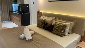 Une chambre d’hôtel avec un lit, des oreillers, une table de chevet, une commode, une œuvre d’art accrochée au mur et, au fond, un coin cuisine.
