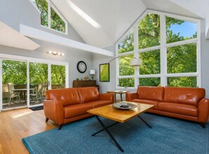 A spacious living room with a large window, a leather sofa, a coffee table, and a blue rug. - Ultimate Treehouse Escape! NEW HOT TUB! Fireplace & Pool Table! (Galena)