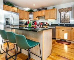 A modern kitchen with wooden cabinets, a stainless steel refrigerator, and a central island with bar stools. - Ultimate Treehouse Escape! NEW HOT TUB! Fireplace & Pool Table! (Galena)