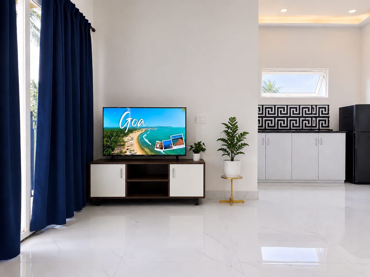 A modern living room with a flat-screen TV displaying a travel ad for Goa, a white cabinet with a gold base, a potted plant, and a kitchen area with a black and white patterned backsplash.