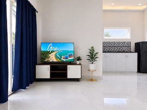 A modern living room with a flat-screen TV displaying a travel ad for Goa, a white cabinet with a gold base, a potted plant, and a kitchen area with a black and white patterned backsplash. - Mogachi Casa Goa Beach Stay (Calangute)