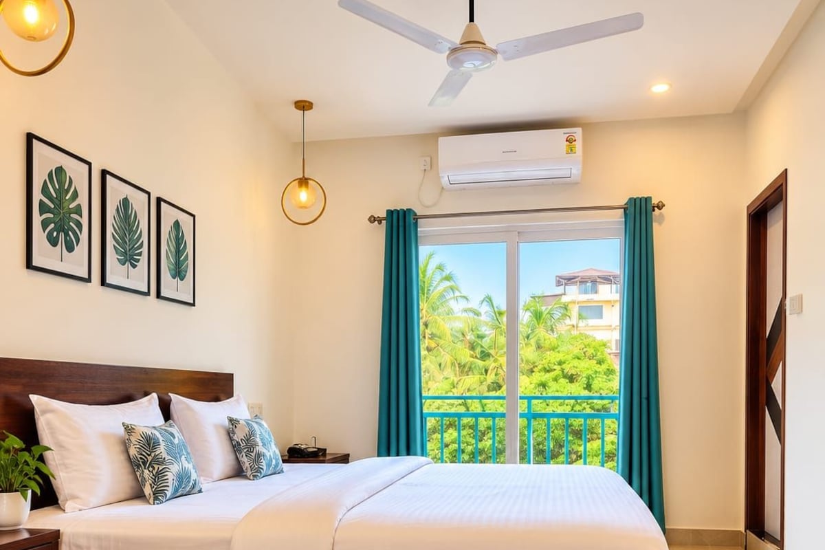 A hotel room with a bed, a nightstand, a ceiling fan, an air conditioning unit, and a view of a balcony with greenery.