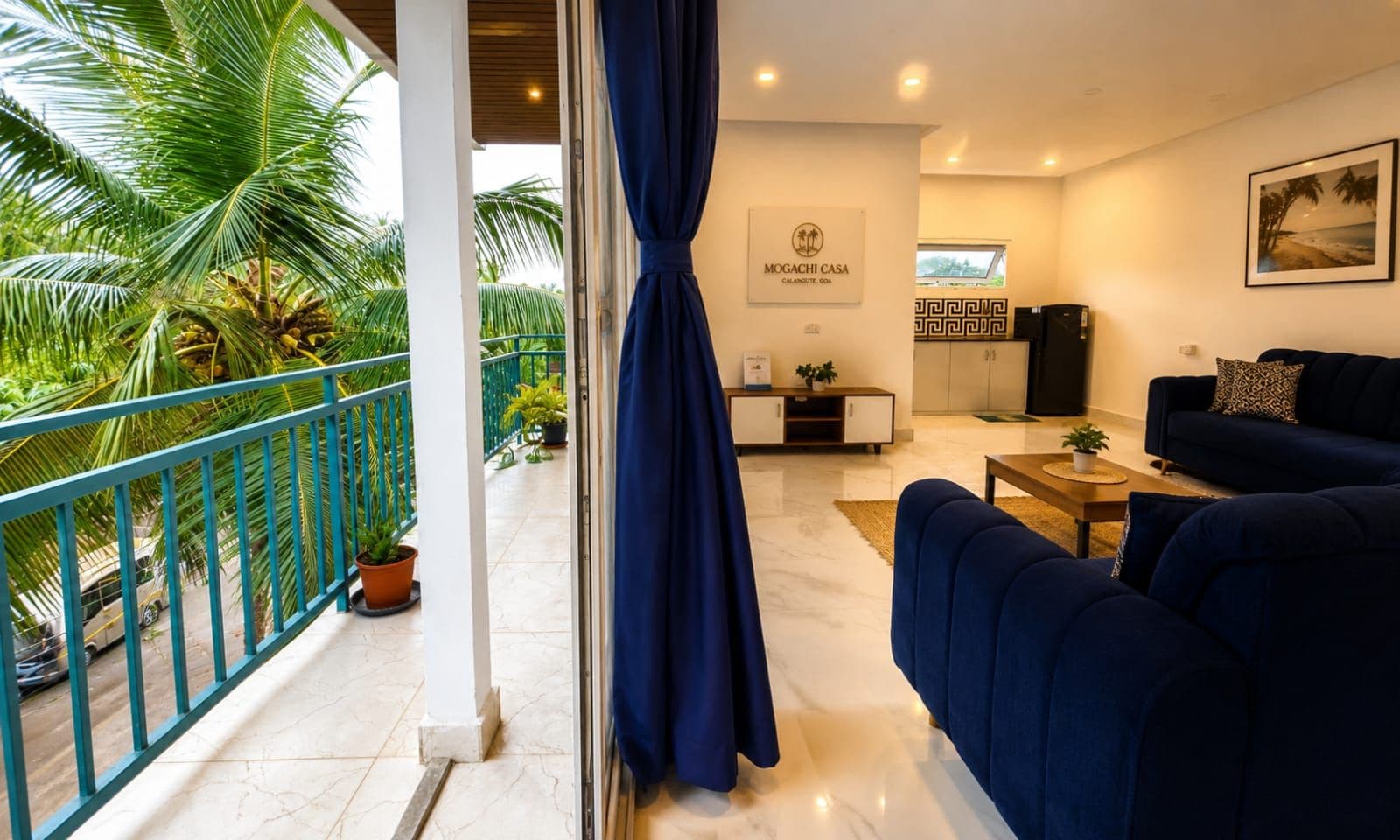 A modern living room with a sofa, a coffee table, and a TV. There is a balcony with a railing and potted plants.