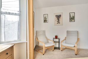 Une pièce comprenant deux chaises de couleur claire, une petite table avec une plante et trois œuvres d’art encadrées accrochées au mur.