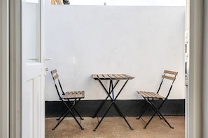 Ein kleiner Sitzbereich im Freien mit Tisch und zwei Stühlen, einer weißen Wand und einer schwarzen Fußleiste.