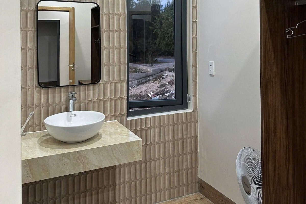 Ein Badezimmer mit großem Fenster, einem Waschbecken inklusive Spiegel und einer Holztür.