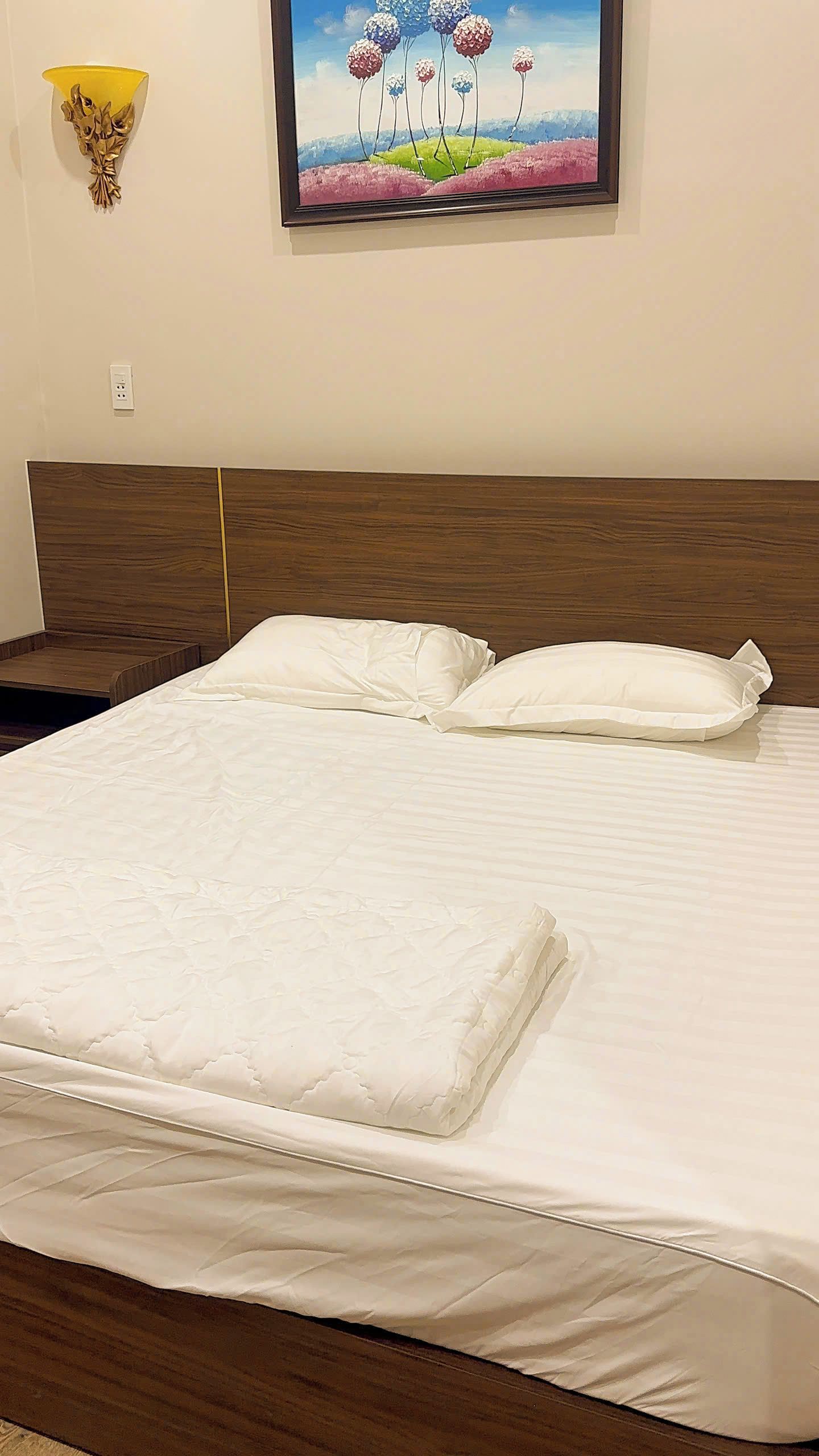 Cama bem arrumada com colchão e travesseiros brancos, cabeceira de madeira e um quadro de flores na parede.