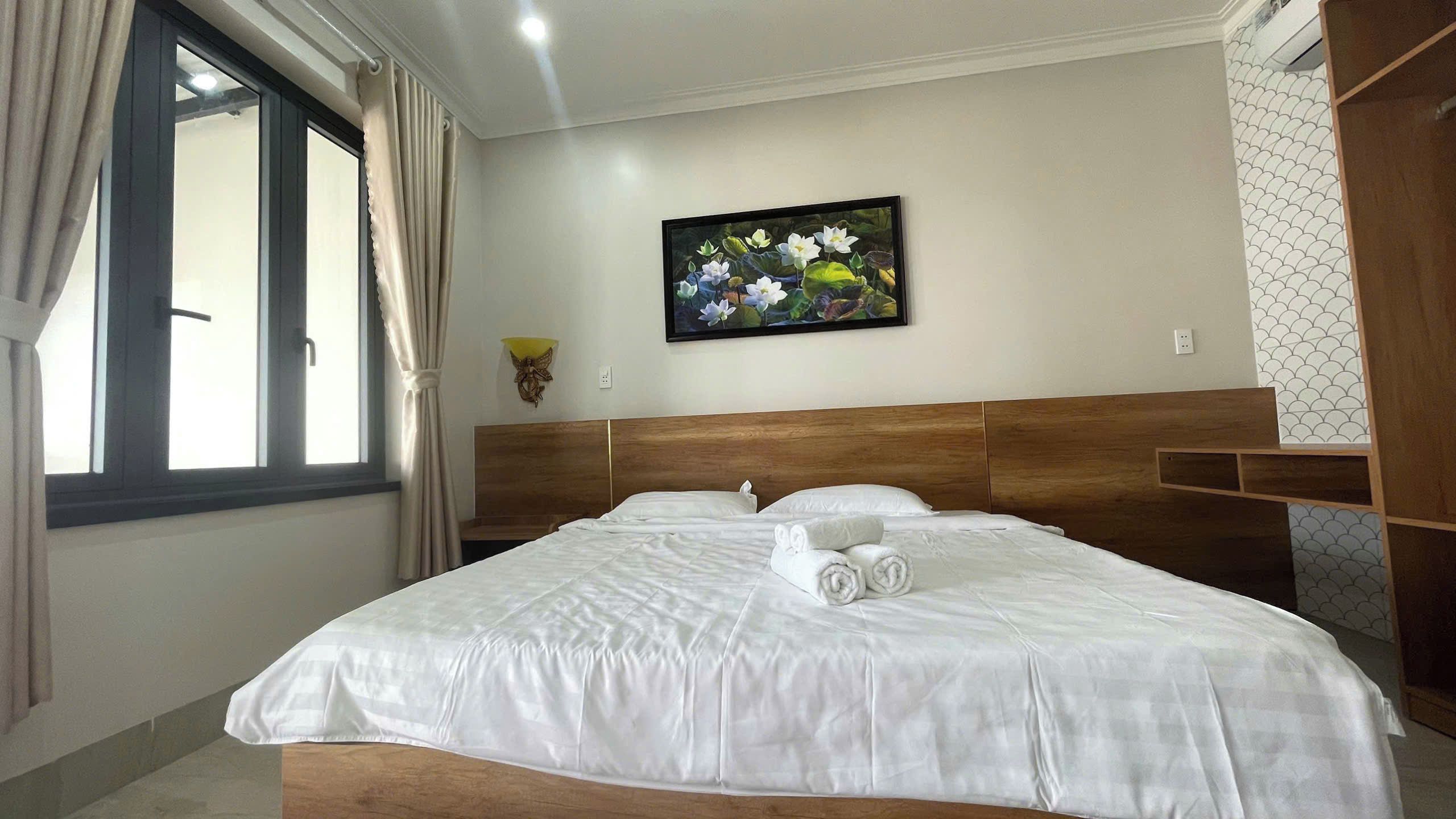 A hotel room with a large bed, a window with curtains, a wall-mounted light, and a framed picture of flowers.