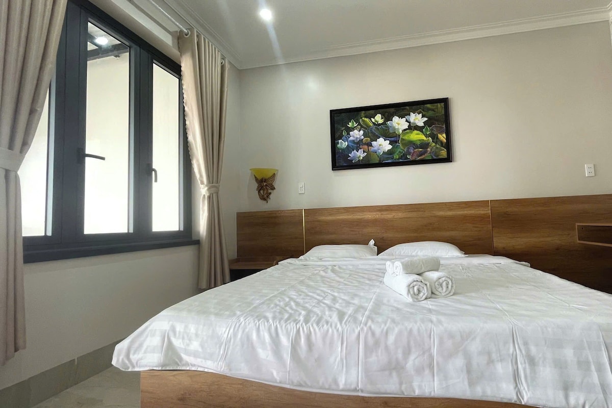 Ein Hotelzimmer mit einem großen Bett, einem Vorhangfenster, einer Wandleuchte und einem Blumenbild in einem Rahmen.