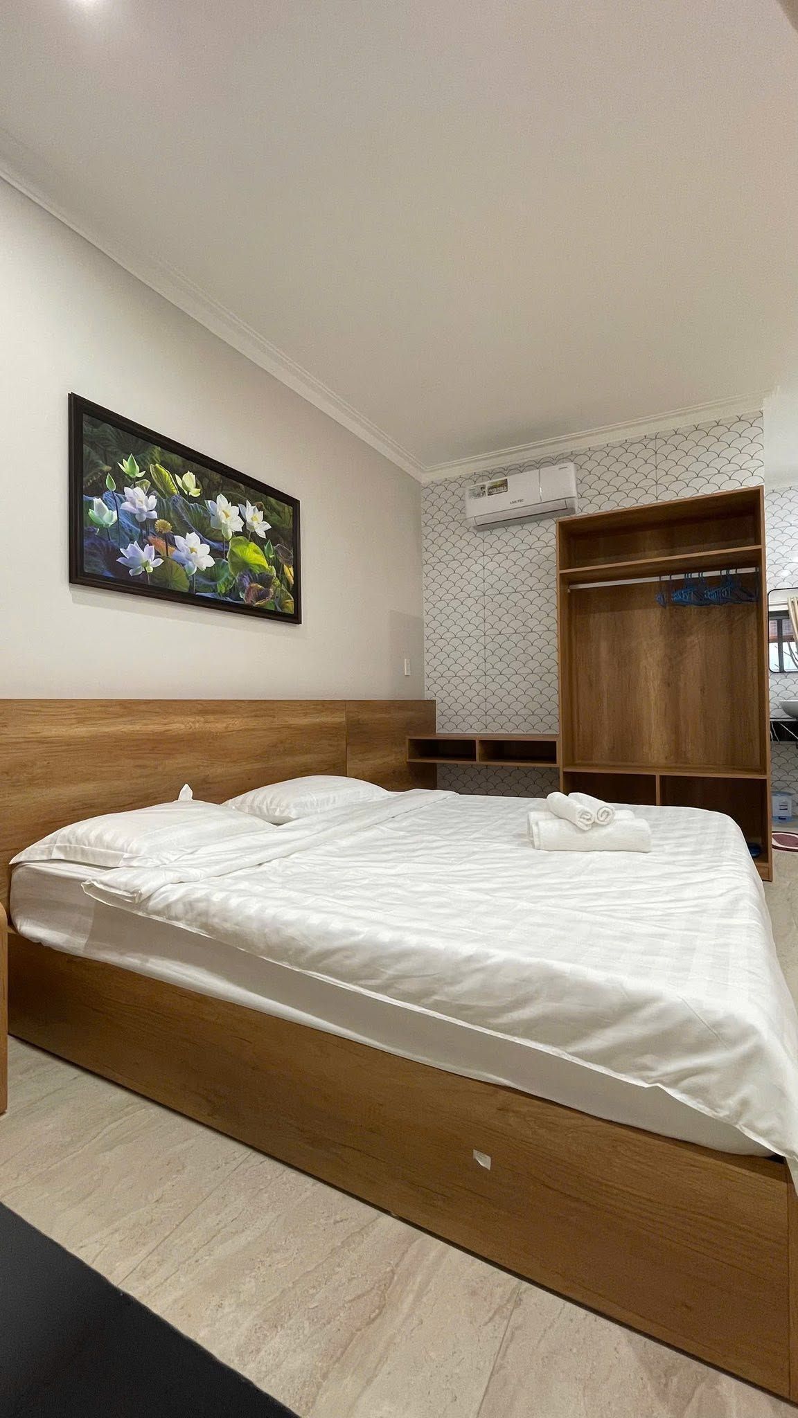 A hotel room with a wooden bed, a painting of flowers on the wall, a wardrobe, and an air conditioning unit.