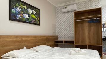 A hotel room with a wooden bed, a painting of flowers on the wall, a wardrobe, and an air conditioning unit.