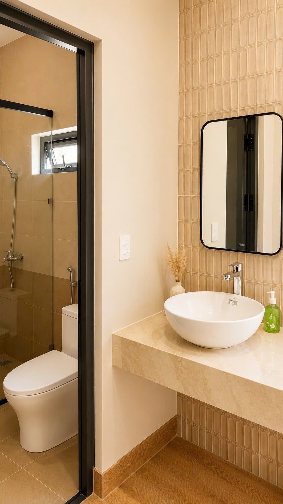 A bathroom with a glass-enclosed shower, a white toilet, a round sink with a mirror above it, and a vase with dried plants on the countertop.